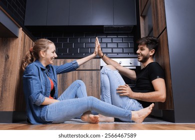 A happy young couple, casually dressed, share a playful high five while sitting on the floor in their modern kitchen. - Powered by Shutterstock