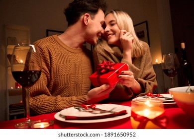 Happy Young Couple Celebrating Anniversary Or Valentines Day Having Romantic Dinner At Home Table. Loving Man Giving Red Gift Box Hugging Beloved Woman Making Present Surprise On Date In Candle Light.