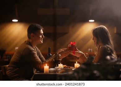 Happy young couple celebrating Valentines day having romantic dinner at home. Loving man giving gift box his woman. Copy space