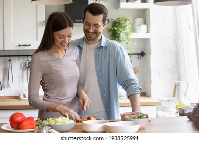 Happy young couple cooking healthy food together, standing in kitchen, smiling woman cutting fresh vegetables for salad, loving husband hugging wife, family enjoying leisure time at home - Powered by Shutterstock