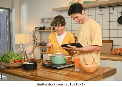 Happy young couple cooking together in a cozy kitchen and following a recipe on digital tablet - Powered by Shutterstock