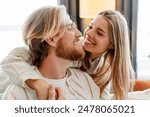 Happy young couple cuddling on the sofa at home, smiling and showing love. Blond woman and bearded man in cozy living room, enjoying each other