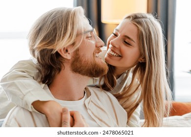 Happy young couple cuddling on the sofa at home, smiling and showing love. Blond woman and bearded man in cozy living room, enjoying each other's company. Joyful and romantic moment captured