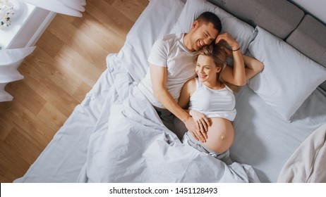 Happy Young Couple Cuddling Together in the Bed, Young Woman is Pregnant and Loving Partner Touches and Caresses Her Belly Tenderly. Sun Shines. Top Down Shot - Powered by Shutterstock