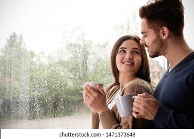 Happy Young Couple With Cups Near Window Indoors On Rainy Day