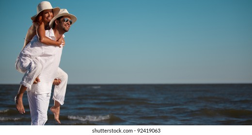 Happy Young Couple Dressed In White, On The Beach, On A Piggy Back Ride