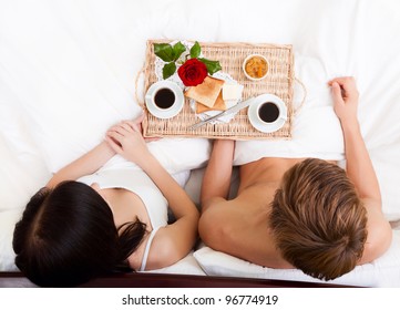 Happy Young Couple Eat Breakfast In Bed In Morning With Red Rose Flower, Lovely Couple Lying In A Bed, Happy Smile Looking At Camera, Concept Of Honeymoon, Valentine Day Top Angle View