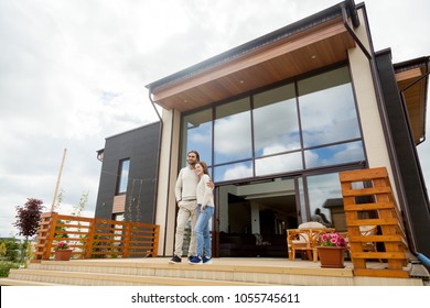 Happy Young Couple Embracing Standing Outside Big House On Front Porch, Smiling Owners Enjoying Luxury Real Estate Wood And Glass Exterior Design, Buying Home, Mortgage Loan, Vacation Rentals Concept