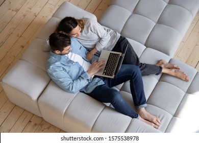 Happy Young Couple Embracing Using Laptop Surfing Internet Relaxing On Comfortable Sofa, Husband And Wife Enjoying Spend Time Online With Computer Device Sitting On Cozy Couch At Home, Above Top View