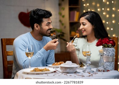 Happy young couple enjoying dinner by talking eachother during candle light dinner at home - concept of romantic moment, dating and togetherness