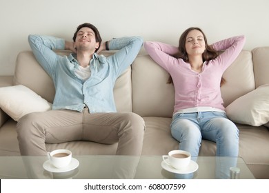 Happy young couple enjoying sitting on sofa at home, resting hands behind the head, relaxing with eyes closed, cup of coffee in the morning, dreaming of vacation, listening to music, leisure lazy day