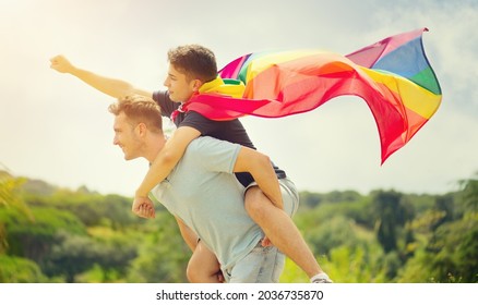 Happy Young Couple, Gay Family With Flying LGBT Rainbow Flag Having Fun Outdoors. Joyful Gay Men Together In A Park. Laughing, Hugging, Enjoying Nature Outside, Over Sunny Sky. Super Hero. Piggyback. 