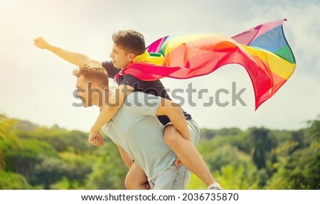 Happy young couple, gay family with flying LGBT rainbow flag having fun outdoors. Joyful gay men together in a park. laughing, hugging, enjoying nature outside, over sunny sky. Super hero. Piggyback. 