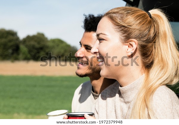 Happy young couple having a coffee break during\
road trip in countryside. Man and woman sitting in car trunk and\
having coffee.