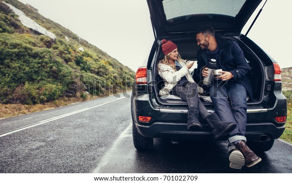 Happy young couple having a coffee break during
road trip in countryside. Man and woman sitting in car trunk and
having coffee.