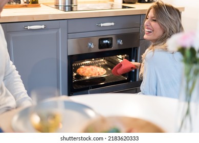 Happy Young Couple Having Dinner At Home And A Young Beautiful Blonde Woman Taking A Pizza Out Of The Oven