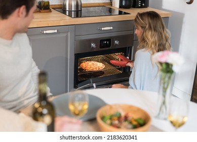 Happy Young Couple Having Dinner At Home And A Young Beautiful Blonde Woman Taking A Pizza Out Of The Oven