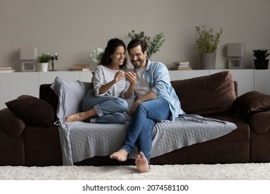 Happy young couple having fun with smartphone at home, sitting on cozy couch, smiling beautiful woman and man in glasses looking at phone screen, watching funny video, surfing internet, chatting - Powered by Shutterstock