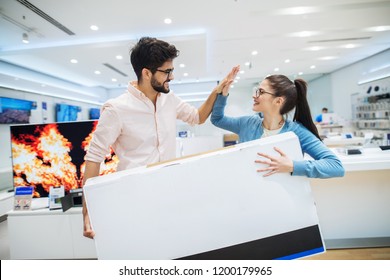 Happy Young Couple Holding Big TV Box In Electronic Store. Smiling And Celebrating Successful Shopping.