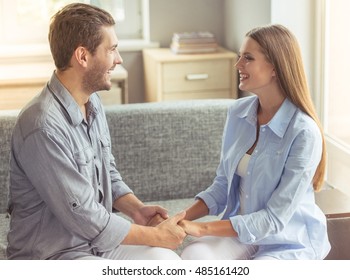 Happy Young Couple Is Holding Hands, Looking At Each Other And Smiling While Sitting On The Couch At Home