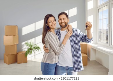 Happy young couple holding a key from a new apartment with cardboard boxes on background, hugging, looking at camera and smiling celebrating moving day. Relocating, real estate, mortgage concept. - Shutterstock ID 2301177321