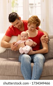 Happy Young Couple Holding Newborn Baby, Sitting On Sofa At Home.