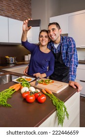 Happy Young Couple In A Home Kitchen Taking A Selfie With A Smarphone While Preparing Healthy Food. Modern Family Lifestyle Concept.