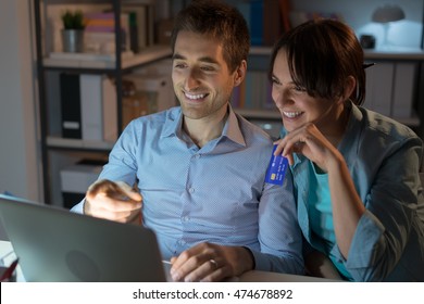 Happy Young Couple At Home Surfing The Web, Networking And Online Shopping Late At Night, She Is Holding A Credit Card