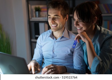 Happy Young Couple At Home Surfing The Web, Networking And Online Shopping Late At Night, She Is Holding A Credit Card