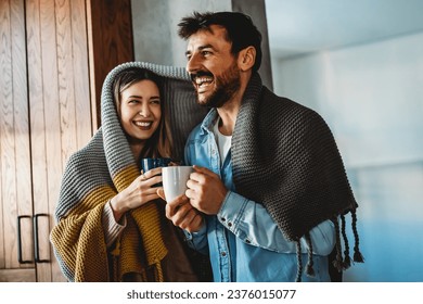 Happy young couple with hot drinks cuddling under warm blanket at home - Powered by Shutterstock