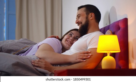 Happy Young Couple Hugging In Bed And Communicating. Side View Of Overweight Girlfriend And Bearded Boyfriend Embracing And Relaxing N Bedroom Together At Home