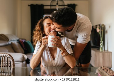 Happy Young Couple Hugging In The Living Room Of Their Home. Happy Couple Kissing Each Other In The Face. Family Having Fun Inside A Comfortable Room Having A Cup Of Coffee Or Tea.