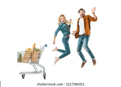 happy young couple jumping near shopping trolley full of products in paper bags isolated on white
