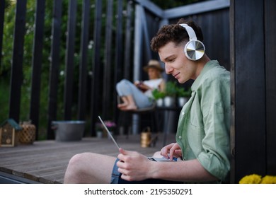 Happy Young Couple With Laptop And Headphones Resting Outdoors In A Tiny House, Weekend Away And Remote Office Concept.