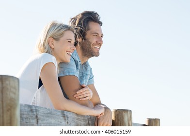 Happy young couple leaning over fence and looking away, copy space. Smiling couple thinking about the future. Young woman embracing her boyfriend outdoor during the sunshine.