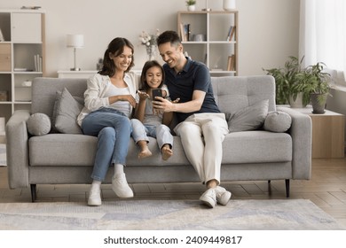 Happy young couple and little kid talking on video call, using smartphone for online family conference communication, resting on comfortable couch together, holding cell, speaking, smiling - Powered by Shutterstock