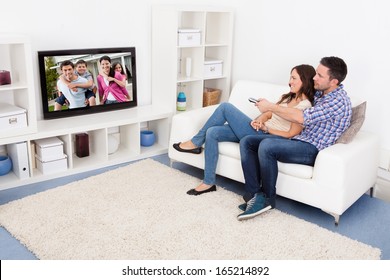 Happy Young Couple In Livingroom Sitting On Couch Watching Television
