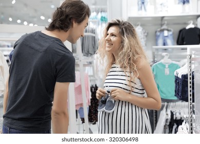 Happy Young Couple Looking For Baby Clothes In A Store.