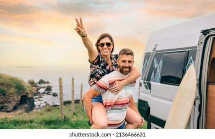 Happy young couple looking camera having fun piggybacking next to their camper van during a trip - Powered by Shutterstock