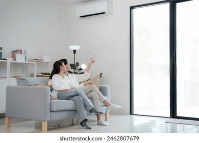 Happy young couple lounges on a sofa in a bright, modern living room, using a remote control to adjust the air conditioning for enhanced home comfort - Powered by Shutterstock