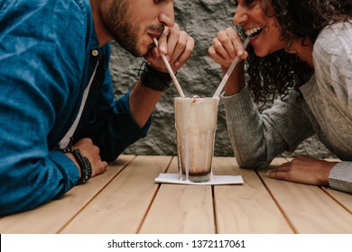 Happy young couple in love at cafe drinking a milkshake from same glass. Young man and woman enjoying a chocolate shake on table.