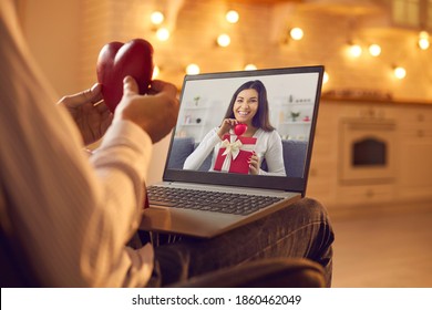 Happy young couple in love having romantic virtual date on Valentine's Day in lockdown. Close-up of man sitting in an armchair at home in the evening, holding red heart and video calling beloved woman - Powered by Shutterstock