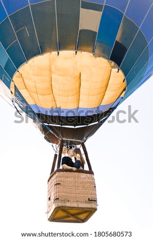 Happy young couple in love kissing in hot air balloon basket, while enjoying their first flight. Romantic travel, engagement, anniversary and honeymoon concept. Bottom view from land