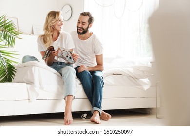 Happy Young Couple In Love Relaxing On A Couch At The Living Room, Reading Magazine, Laughing