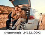 A happy young couple in love sitting in the open trunk of a car on a warm blanket, drinking tea from a thermos and taking a selfie on a smartphone on their weekend. Travel and vacation concept.