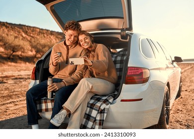 A happy young couple in love sitting in the open trunk of a car on a warm blanket, drinking tea from a thermos and taking a selfie on a smartphone on their weekend. Travel and vacation concept.