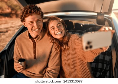 A happy young couple in love sitting in the open trunk of a car on a warm blanket, drinking tea from a thermos and taking a selfie on a smartphone on their weekend. Travel and vacation concept. - Powered by Shutterstock