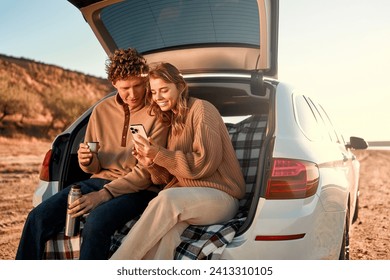 A happy young couple in love sitting in the open trunk of a car on a warm blanket, drinking tea from a thermos and using a smartphone on their weekend. Travel and vacation concept.