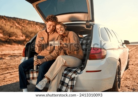 A happy young couple in love sitting in the open trunk of a car on a warm blanket, drinking tea from a thermos and taking a selfie on a smartphone on their weekend. Travel and vacation concept.