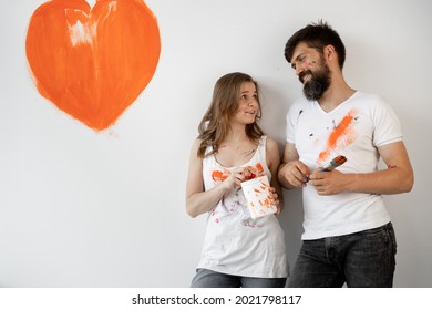 Happy Young Couple In Love In White T-shirts Making Renovations, Updating The Painting Of The Walls, Getting Ready To Move To A New Home, Painted An Orange Heart On The Wall. Copy Space.